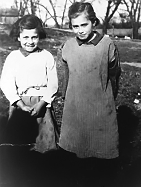 Black and white photograph: Two female children standing beside one another outside. They appear to be smiling. One is wearing a long skirt and top and the other a short-sleeve blouse with a basic work dress over the top.