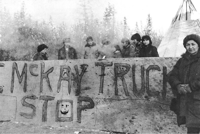 The black-and-white image shows a group of people standing behind a large, hand-painted sign in an outdoor setting. The sign has the words MCKAY TRUCK STOP written in uneven, capital letters. Two smiley faces are drawn within the letters. The people appear to be wearing warm clothing, indicating a cold weather environment. In the background, there is a blurred view of trees, suggesting a forested area. On the right side, a person is standing in front of a teepee. The overall atmosphere seems informal and community-oriented.