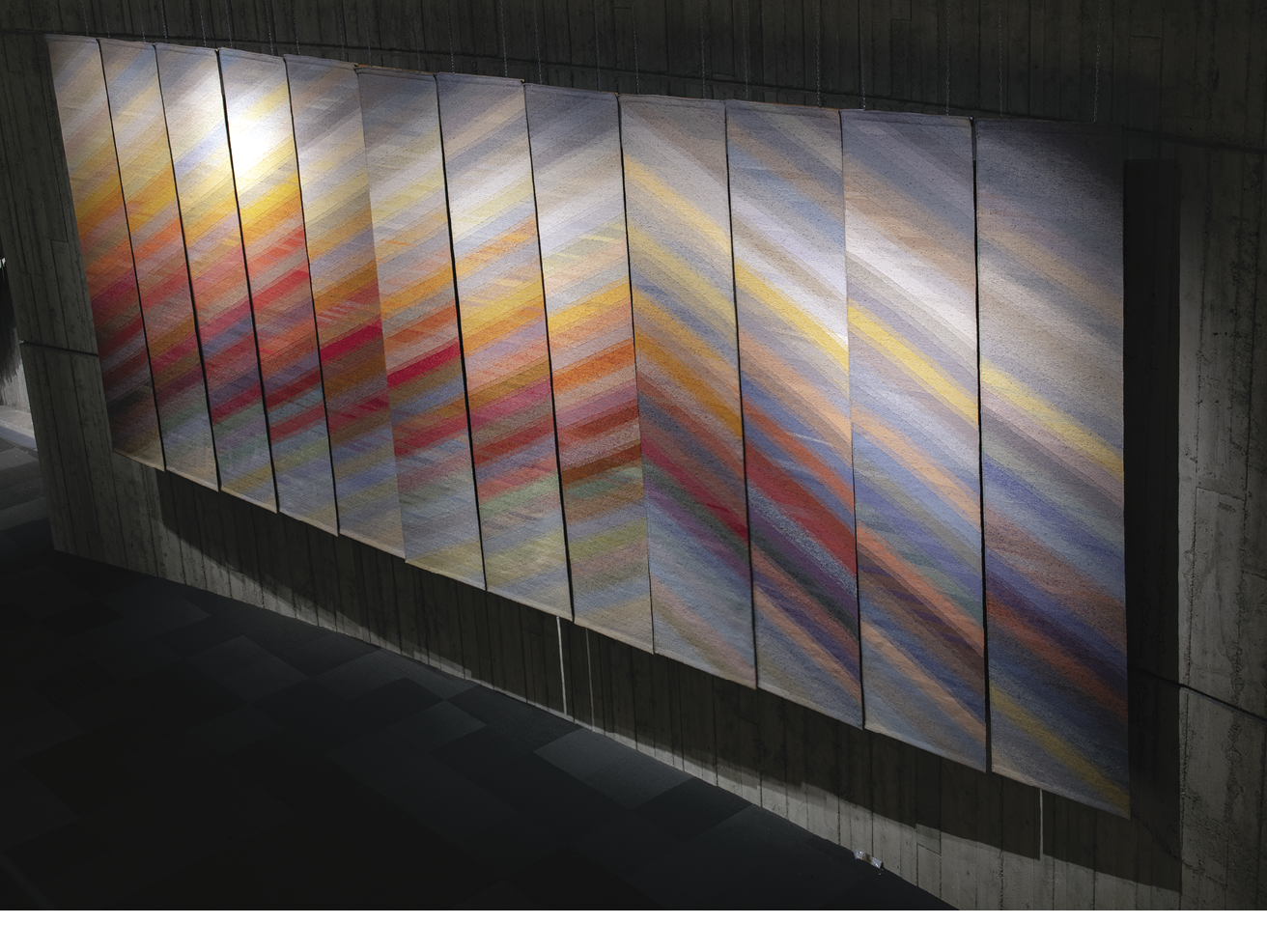 Photo: looking down at a series of twelve tall hangings that from a pyramid pattern from orange, red, pin, and yellow lines against a blue-grey background. 