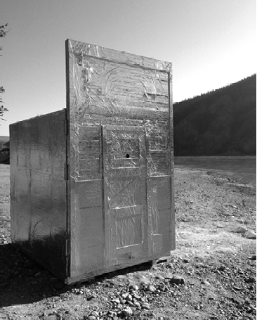 Black and white photograph, a box covered in reflective foil sits on a rocky riverside, the box has a large door with a small peephole at the front.