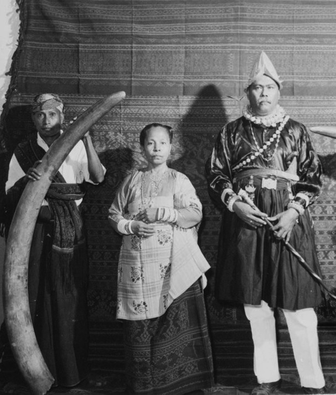 Black and white photograph. 3 opulently dressed people stand in front of a decorated cloth wall. From left to right, a man wearing a sarong, sash, shirt and headband holds an elephant tusk as tall as him. A woman stands in the middle wearing a light coloured top and decorated dark skirt. A tall man wearing a Portuguese morion helmet and a shiny shirt, stands facing forward, holding a cane.