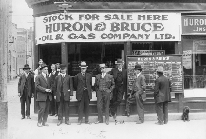 Figure 5-6 “Investors waiting to buy oil and gas stocks outside Huron and Bruce Oil and Gas Company, Calgary, Alberta.”  Black and white photograph of 11 men and dog outside the Huron and Bruce Oil and Gas Company at the corner of 7th Avenue and Centre Street. On the right hand side of the picture is a blackboard listing the stock information for several different offerings. A hand-written message at the bottom of the picture identifies the location as “7th Ave & Centre” while a not under the man in a three piece suit leaning up against the blackboard says “Dad.”