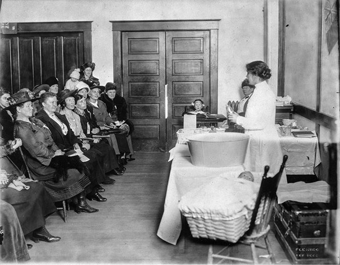 Black and white photograph: An Alberta public health nurse in a white dress on the right side of the photograph is speaking before a seated group of women on the left during a child welfare clinic. In the foreground of the image, there is a metal baby’s bathtub and a chair with a model baby in a basket on it. Other model children and child-raising props are distributed on guests around the speaker.