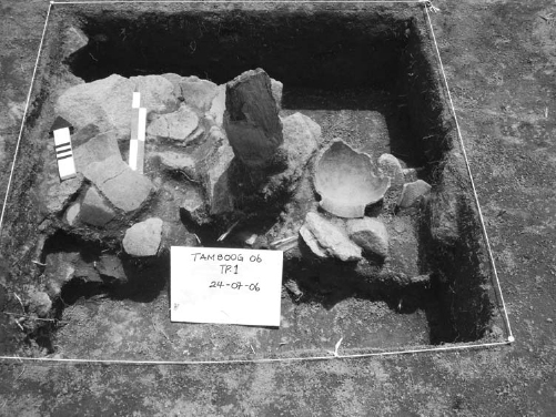 A standing stone is surrounded by a cluster of broken pots at its base. Text on a card placed on the site reads, TAMBOOG 06 TP.1, 24-07-06.