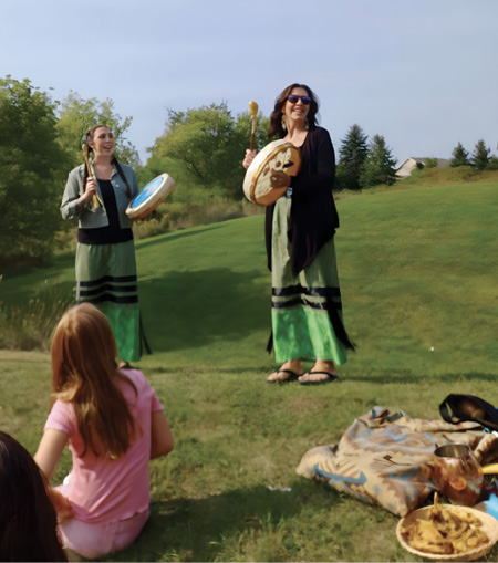 Colour photograph. Two women are standing in a field, playing hand drums. They are wearing green and black ribbon skirts. A young red-haired girl wearing pink is sitting in the foreground, with her back to the camera, and there are colourful drum bags and a bowl of what appears to be Indigenous sacred medicine to her right.