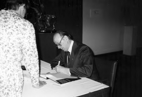 A photograph shows two-person, one of them is sitting on a chair and writing on a book while the other looking at him. A wall with some design can be seen in the background.