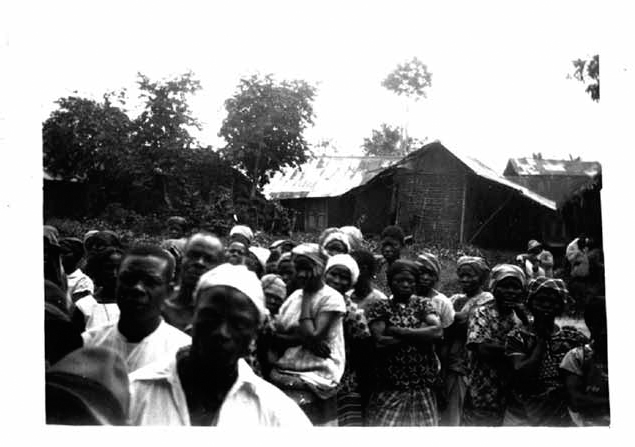 A black and white photograph shows villagers gathered in front of a building.