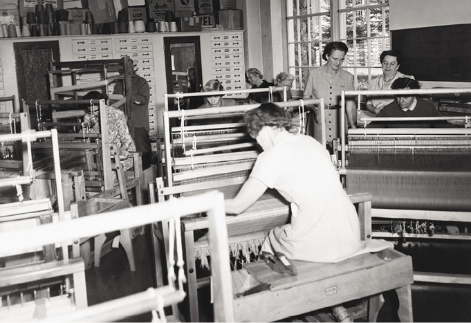 Photo noir et blanc : une femme de dos en train de tisser dans une salle de classe où d’autres femmes travaillent, sous le regard d’autres femmes. 