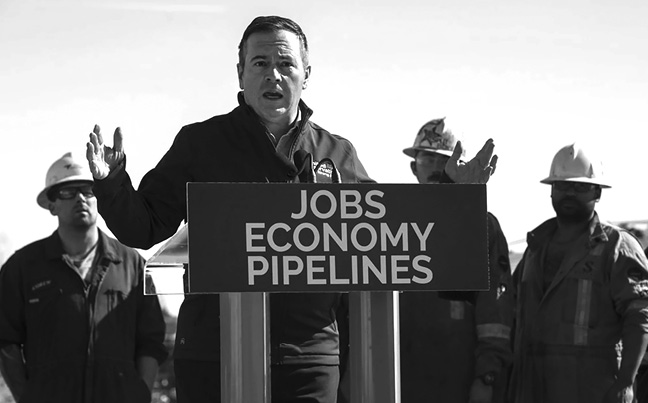 Jason Kenney stands outside in front of a podium that says “JOBS ECONOMY PIPELINES.” He is speaking and has both hands raised. Behind him stand three men wearing hardhats and coveralls.