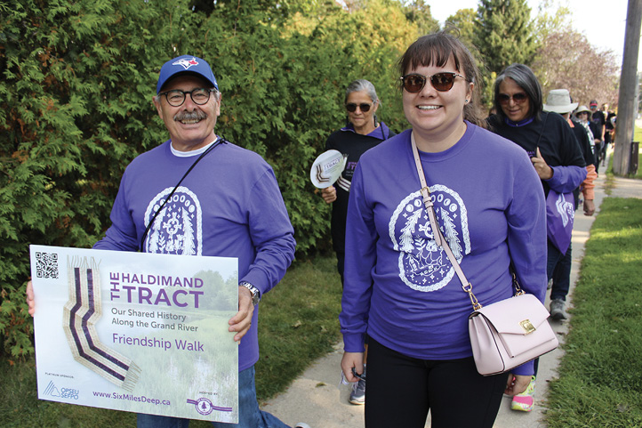 Colour photograph. A group of people are walking along a suburban footpath. At the front are a man and a woman wearing purple shirts with white Indigenous motifs in a circle on the chest. The man is smiling, wearing glasses and a cap, and holding a sign saying “The Haldimand Tract Friendship Walk”. The woman wears sunglasses, has bangs, and has a pink shoulder purse. There is a large hedge next to the group, and the people in the background are talking.
