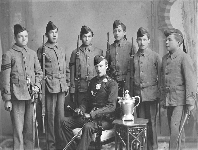 Black and white photograph: Formal professional photograph in what appears to be a studio setting of six male Indigenous students in their cadet uniforms with firearms, standing behind a male non-Indigenous teacher who is sitting in a chair. Beside their teacher, a trophy sits on a plinth.