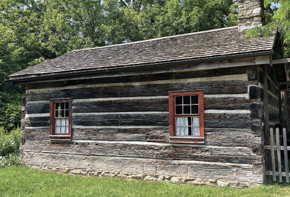 Colour photograph. A log cabin, with two glass windows and shingled roof. There is a stone chimney, and a fence on the right side. There are mature trees behind the cabin.