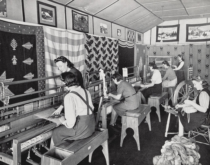 Photo noir et blanc : cinq femmes le long d’un mur drapé de textiles, assises sur des bancs en train de tisser. Une autre femme observe, en montrant les fils du métier à tisser de la première femme, tandis qu’une autre femme est assise à un rouet. 