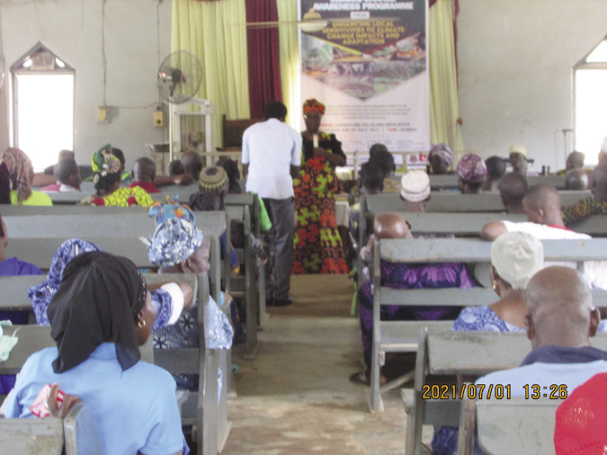 Colour photograph: About 28 workshop participants (women and men) sit on benches; a woman in African dress leads the session, with a poster projected on the wall behind her.