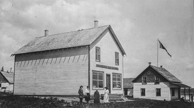 The photograph portrays a historical scene featuring two main wooden buildings set in a grassy area under a cloudy sky. In the foreground, the larger building is a simple, two-story structure with a pitched roof and two visible windows on the second floor. The facade has a sign above the main entrance reading HUDSON'S BAY STORE. The building's exterior is made of horizontal wooden planks. Four people—three women and one man—stand in front, wearing early 20th-century attire. To the right is a smaller building with a flagpole displaying a tattered flag. This building also has a wooden structure, with several windows and a small porch. The background includes some trees and more wooden structures.