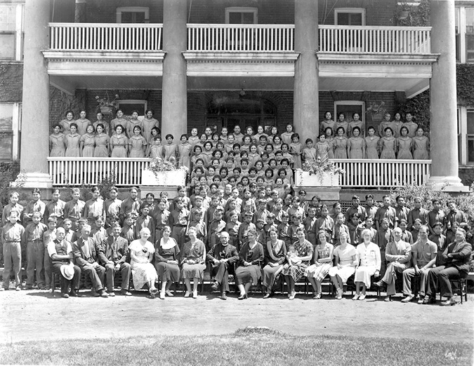 Black and white photograph: Group of approximately 100 staff and students of the Mohawk Institute.  The staff are seated in rows on the lawn outside the front of the school and the students are standing in rows behind them, going all the way up the steps to the front door and across the front porch.  The children are wearing uniforms and are separated by gender.  The students range in age from what appears to be young children to teenagers.