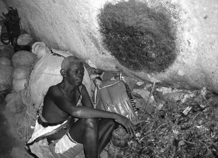 A man sits inside a rock shelter. A rock above him has a big burnt spot. Below the spot are a heap of used ritual equipment and residues.