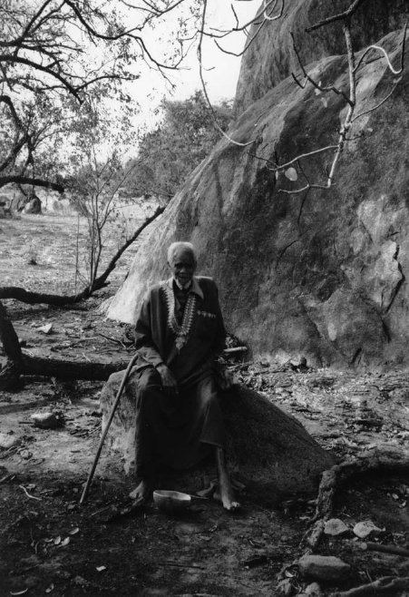 An elderly man is seated on a rock. A stick and a bowl lie in front.