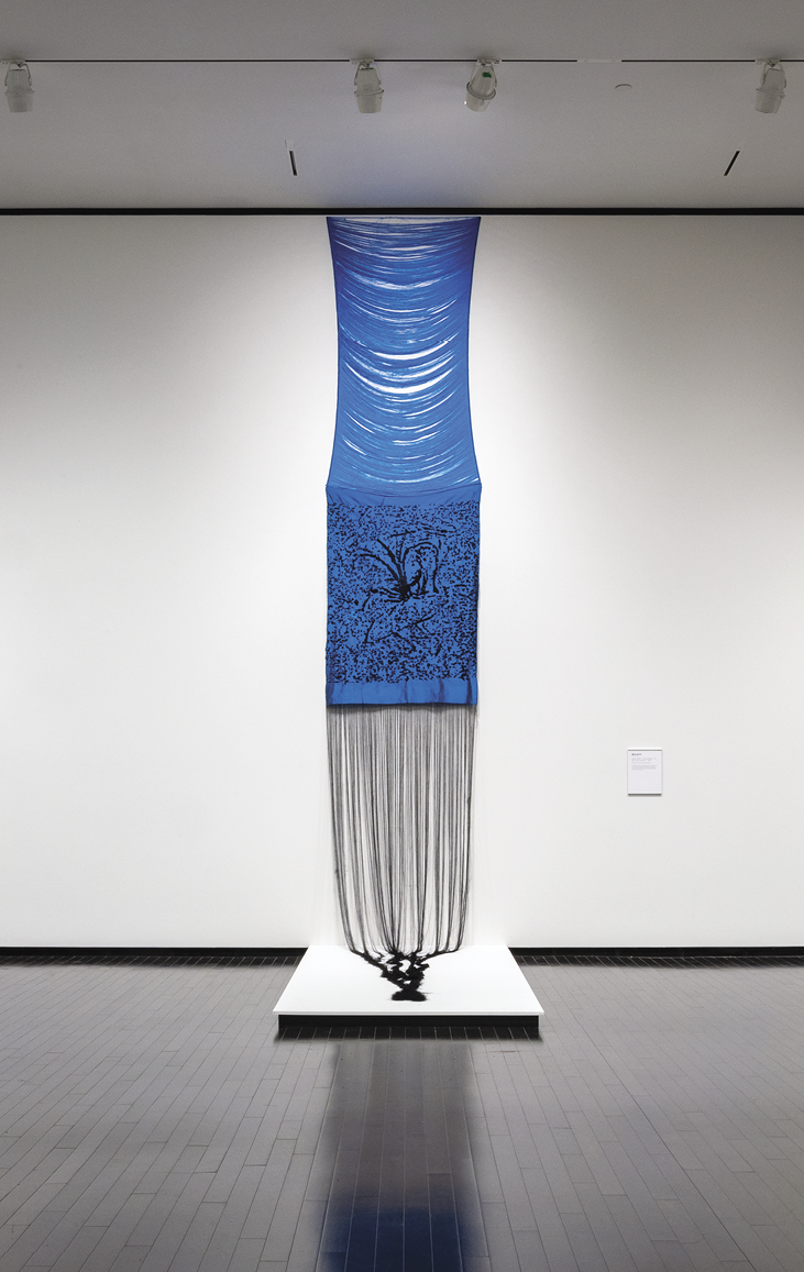 Photo: a tall blue artwork. The top third is horizontal threads bowed in the middle, the middle third is a blue and black swirl design, the bottom third is a long black fringe that pours out onto a white shelf at the bottom. 