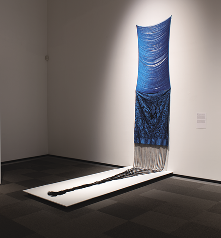 Photo : une grande œuvre d’art bleue. Le tiers supérieur est constitué de fils horizontaux courbés au milieu, le tiers central est un motif tourbillonnant bleu et noir, le tiers inférieur est une longue frange noire qui se déverse sur une étagère blanche dans la partie inférieure de l’œuvre d’art.
