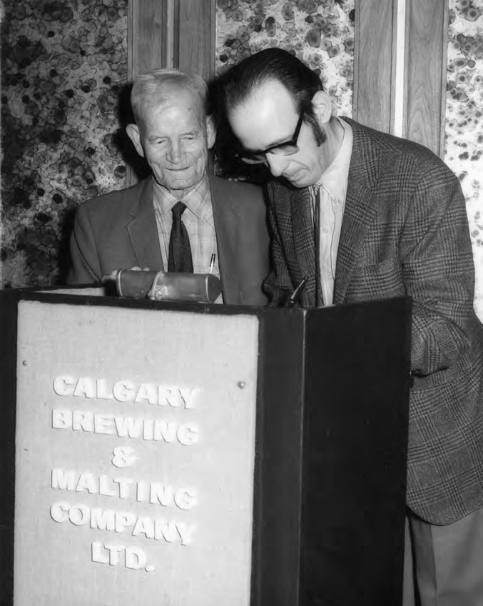 A photograph of two people in formal uniform, one is old while the other is young and a podium with the text CALGARY BREWING and MALTING COMPANY LTD. A background with a flower design can be seen.