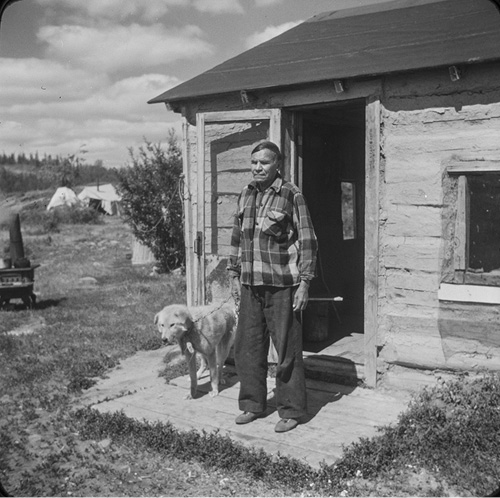 Black and white photograph: Jonas Laviolette stands on the front step of his single-story wood cabin on a cloudy day, wearing a plaid shirt and black pants. He has a large, white long-haired dog on a lead to his right. A black wood stove is in the foreground, and two white tents are in the background.
