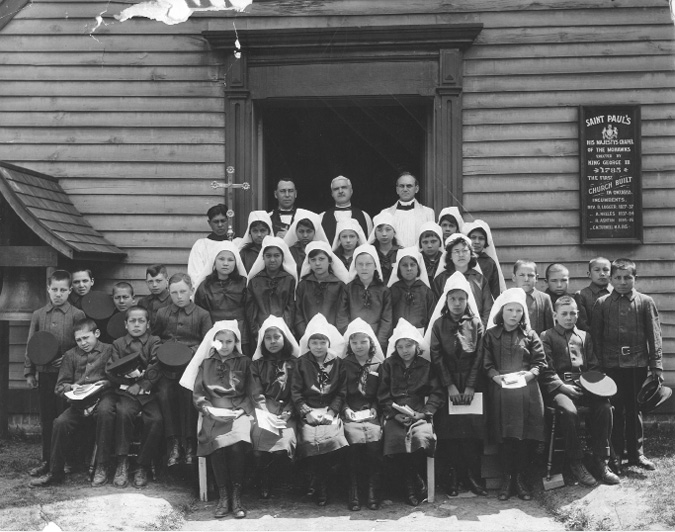 Black and white photograph: Three rows of children outside the front of the Mohawk Chapel, with a row of priests in religious garb standing behind them. The female students are in uniforms and have scarf-like covers on their hair.  They are sitting and standing in the middle of the rows. Boys in uniforms are located on both sides of the girls. Many of the children are holding papers and what appear to be bibles.