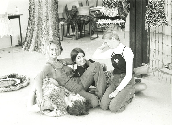Black and white Photo: Three smiling young women leaning against a man on the floor. They are in casual 1970s dress.