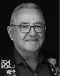 Black and white photograph: Portrait of a man wearing glasses and smiling, wearing a collared shirt with floral decorated garment on top.