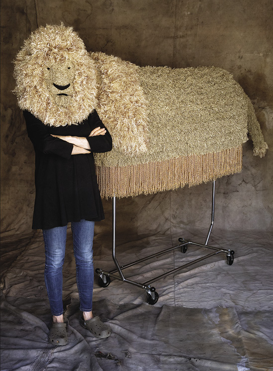 Photo: a person in usual clothing but wearing a shaggy lion’s head stand in front of the textile body of the lion on a metal frame, with a long fringe. 