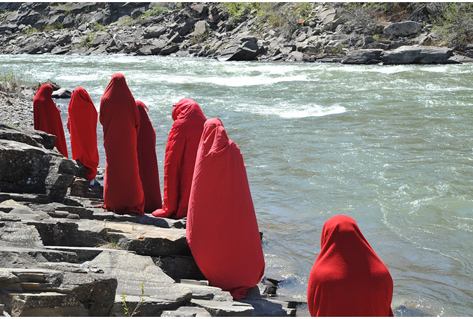Along the boulders of a rocky river shoreline, in the foreground of the image is a row of people wrapped in red fabric. The view is of the peoples’ backs, creating a line of red figures along the horizontal line of the shore.