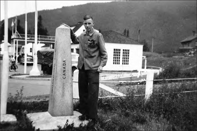 A photograph shows a man standing beside a pillar with Canada written over it. A house, trees and mountains can be seen in the background.
