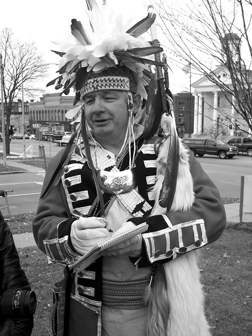 Black and white photograph: Adult Indigenous male standing outside wearing a traditional headdress taking notes in a book.