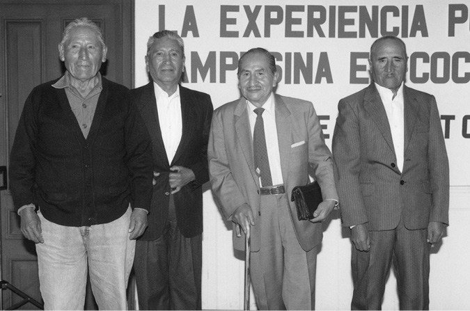 Four older men pose on the podium of a convention centre. The first on the left gazes slightly off to the side and wears sweater and jeans. The other three have a frontal gaze and wear suits, although only the third man in the row stylishly wears a tie and pocket square, leans on a cane, and carries a handbag.