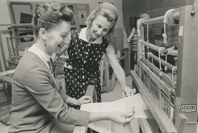 Black and white Photo: a middle-aged woman works at a loom while another in a black dress with white diamond pattern looks on. 