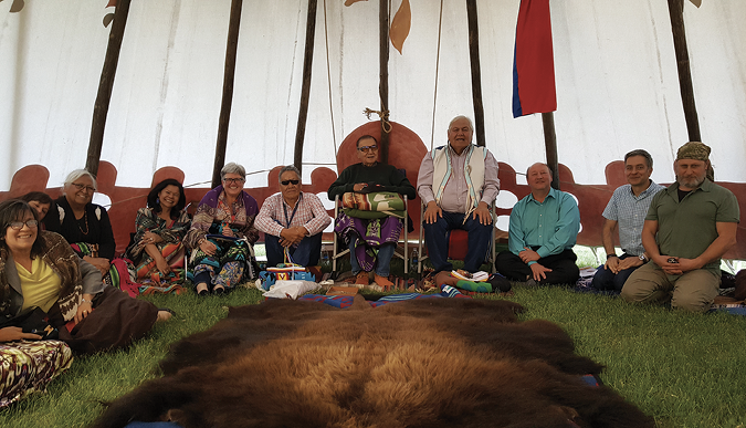 Colour Photograph: A group of elders and others seated in a circle inside a tipi. A buffalo hide is lain out in front of them, with a variety of cultural objects on it. 