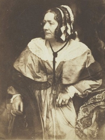 Black and white photograph. Portrait of an older white woman, seated on a chair, with a serious expression. She is wearing a dress over a white blouse with lace collar, and a black bonnet. She is holding eyeglasses in her hand and looking off to the left.