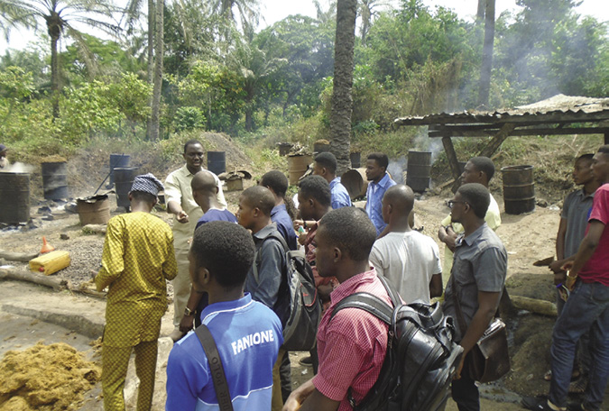 Colour photograph: A group of about 15 young people, mostly male, stands outside listening to a male teacher.  Fires in barrels are smoking in the background, and concrete slopes on the ground may be areas where food is processed.