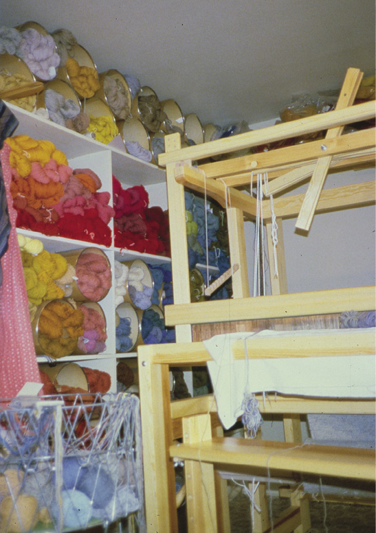 Photo: The many frames of a loom sit in a room next to shelves containing many different colours of yarn in balls. 