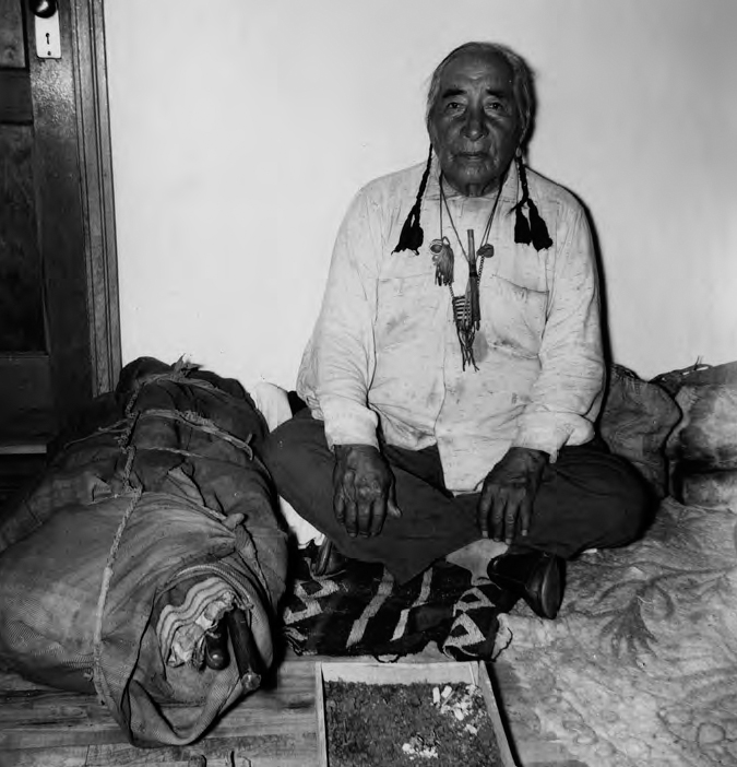 A photograph shows an old man sitting on the floor. He is wearing a white shirt and black pants. A door and wall can be seen in the background.