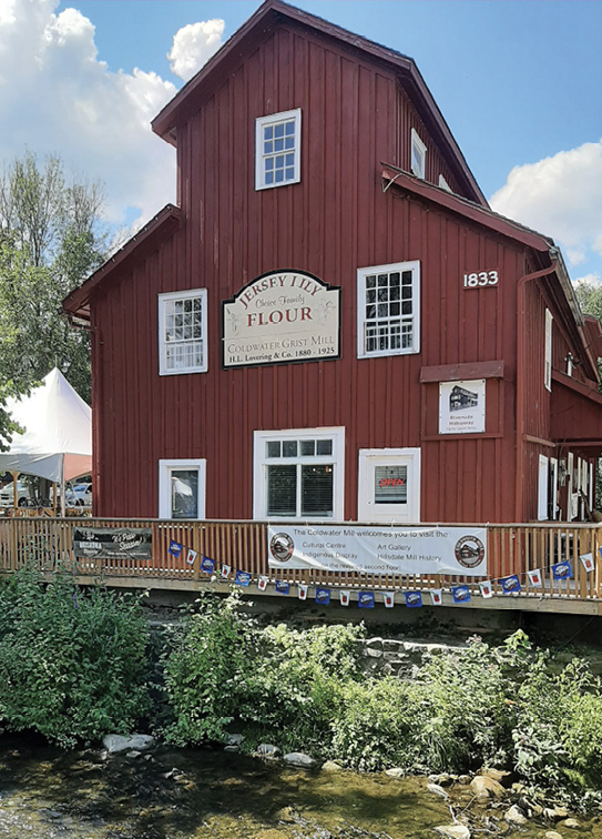 Colour photograph. A red wooden mill building, with signage showing it is now a museum. There is a patio with a fence and banners on it. The mill has white framed windows and a door, and is three stories high. There is a small creek with bushes on the shoreline in front of it.