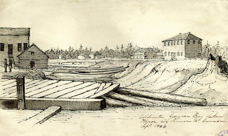 Black and white drawing. A town scene, with a large wooden platform (perhaps a corduroy road of wooden planking) or a dock in the foreground. There is a river behind this, with a large wooden building on the left bank, with two people standing in front of it. The right bank rises steeply, and there are several large wooden buildings along this, visible into the distance. There is forest behind these. A signature and caption are written at the bottom of the drawing.