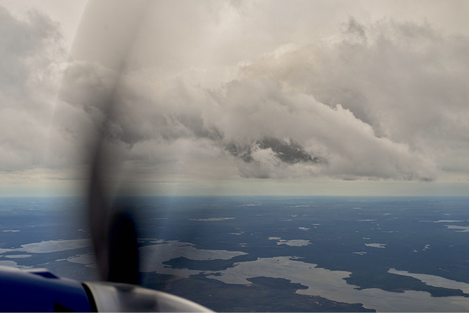 An aerial view from a small plane, the propeller is creating a wave image of the stretch of land, green forested areas and waters below. Cloud ad blue sky sit at the top of the image.
