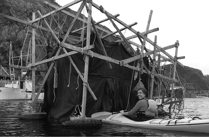 Black and white photograph, a woman in a kayak floats into a floating wooden scaffold; this has a curtained area hanging inside it; boats and houses sit in the background, and cliffs rise close behind at the water’s edge.