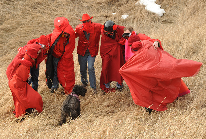 Ten people, wrapped in red fabric, form a semi-circle in tall grasses. In the middle of the semi circle is a black dog. Wind is blowing wildly and the red fabric of one in the circle is stretched like a flag.