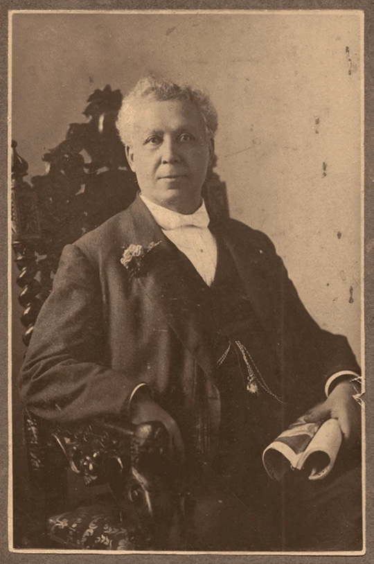 Sepia historical photograph. Portrait of a man seated in an elaborately carved high-backed wooden chair. He has curly greying hair, and faces the camera. He is wearing a suit with a floral decoration pinned to his lapel, and is holding some papers in his left hand. His right arm rests on one of the carved arms of the chair.