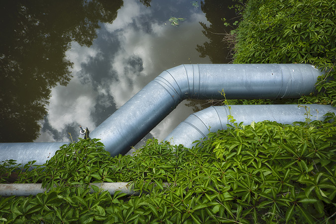 Two large steel aqueduct pipes sit one on top of the other stretching horizontally across a reflective surface of water. Green vines are growing from the shore and trailing up between the two pipes. The reflections of trees anchor each side of the view with a blue sky and wispy white clouds above. The photographer stood on a bridge over the Lahn River in Marburg, Germany.