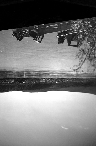 Black and white photograph, an upside-down view of a lake, speedboats and bushes are seen in the foreground, houses and forested hills rise in the background at the far lakeshore.