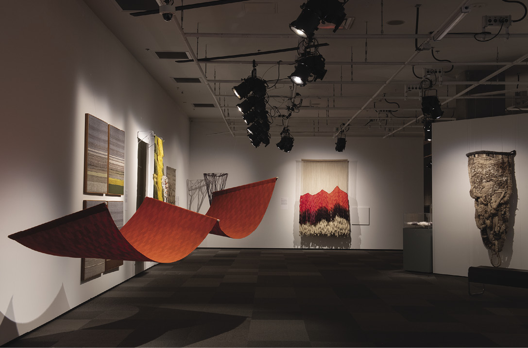 Photo: a dark gallery with a spotlighted artwork including a wavey red-orange carpet, a red-brown-tan-grey hanging where the red imitates the shape of mountains, and a piece that is almost mask-shaped and id complex, bumpy, interwoven.