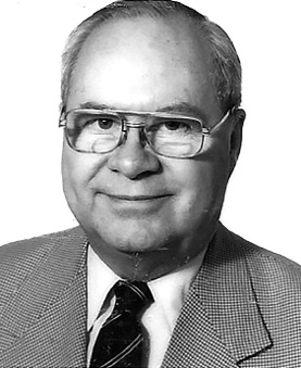 Black and white photograph: A portrait of a man wearing glasses, a suit jacket, a white shirt, and a dark tie. He is smiling gently and looking directly at the camera.